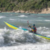 Kayaker wearing a teal helmet and red life vest paddling a bright yellow-green kayak through white waves near a rocky shoreline.