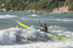 Kayaker wearing a teal helmet and red life vest paddling a bright yellow-green kayak through white waves near a rocky shoreline.