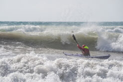 Person in a kayak paddling through large ocean waves, wearing a red jacket and neon green hood, with spray around them