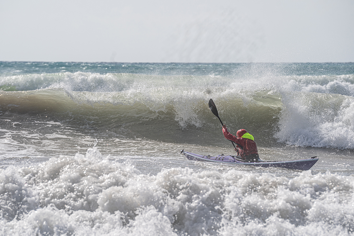 Person in a kayak paddling through large ocean waves, wearing a red jacket and neon green hood, with spray around them