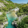 A turquoise river winds through a rocky, forested canyon with kayakers paddling along the water under a bright blue sky.