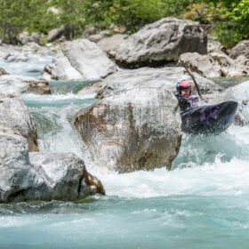 Person in a purple kayak navigating turquoise rapids among gray rocks, wearing a pink helmet.