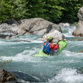 Person paddling a bright green kayak through white-water rapids, wearing a red jacket and yellow helmet in a rocky river canyon