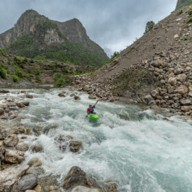 Kayaker in a bright green boat paddles through whitewater rapids in a rocky canyon with steep hills and mountains in the background.