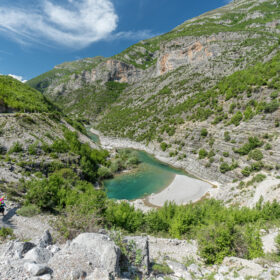 A hiker in a colorful outfit walks along a rocky trail beside a turquoise river cutting through a steep, green-covered canyon with a small sandy beach in the foreground.