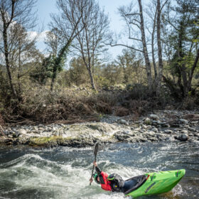 A person in a bright green kayak paddles through whitewater along a rocky riverbank with leafless trees in the background, wearing a red jacket and helmet.