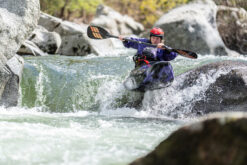 Person wearing a red helmet kayaks through whitewater rapids between rocks and boulders in a narrow river route.