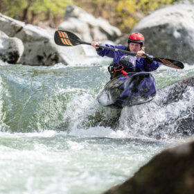 Person wearing a red helmet kayaks through whitewater rapids between rocks and boulders in a narrow river route.