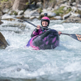 Person wearing a pink helmet and purple wetsuit paddling a kayak through whitewater, GoPro mounted on helmet.