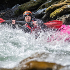 Person whitewater kayaking in a pink kayak, navigating splashing waves with a red Werner paddle and helmet on rocky rapids surface