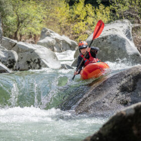 Person in a red kayak navigating fast, rocky whitewater with paddles raised amid large boulders.