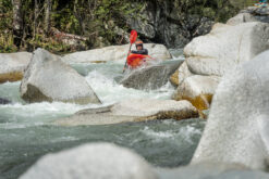Kayaker navigating a rocky whitewater river in a red kayak with a paddle raised over rapids.