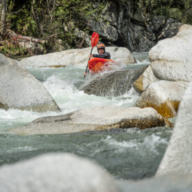 Kayaker navigating a rocky whitewater river in a red kayak with a paddle raised over rapids.