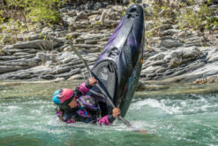 Person in a pink helmet and purple gear trying to upright a purple jet ski that’s tipping in a rocky river.