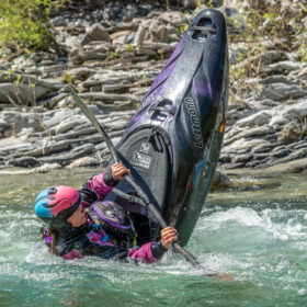 Person in a pink helmet and purple gear trying to upright a purple jet ski that’s tipping in a rocky river.