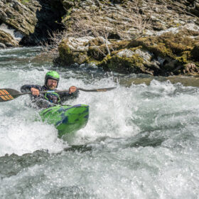 Kayaker in a bright green boat paddling through whitewater rapids, wearing a green helmet near a rocky riverbank.