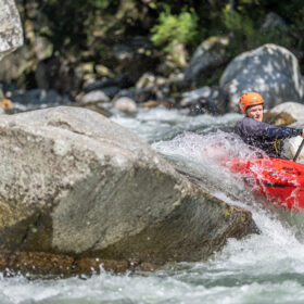 Person whitewater kayaking in a red kayak around large rocks in a rapid, wearing an orange helmet and dark wetsuit