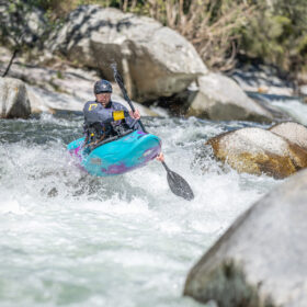 Person in a turquoise kayak paddling through a rocky river rapid, splashing water around.