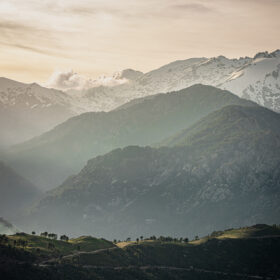 Snow-capped mountains at dawn with hazy green valleys and a soft orange sky.