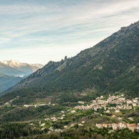 Mountain valley with a small village tucked into the hillside, framed by forested peaks under a blue sky.