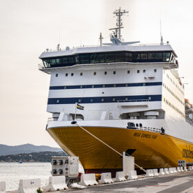 Large white passenger ferry with a yellow bow docked at the harbor, calm water in the background.
