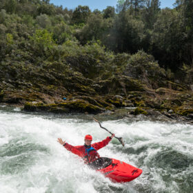 Kayaker in red gear navigating whitewater rapids in a red kayak beside a rocky, forested riverbank.