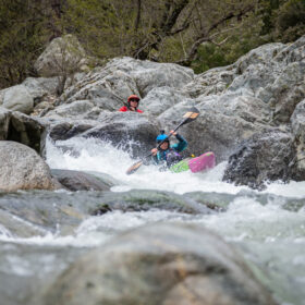 Two kayakers paddling through rocky white-water rapids, wearing helmets and life jackets, between large boulders.