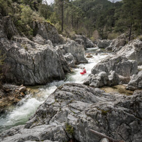 Person in a red kayak paddling through a rocky river canyon lined with pine trees.
