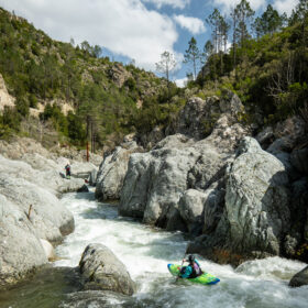 Two kayakers navigate a fast, rocky river through a canyon with pine-covered hills.