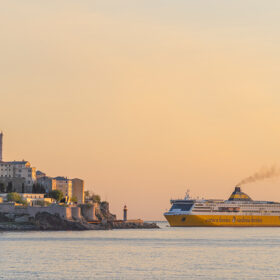 Sunset over a hillside town with a lighthouse, while a yellow Corsica Ferries ship sails nearby.