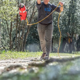 Person walking toward the camera on a dirt trail in a wooded area, holding an orange rope with a red bag attached.