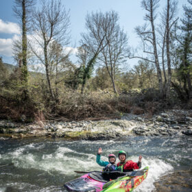 Two people wearing helmets kayak together in a colorful tandem boat on a choppy river, waving at the camera with trees and blue sky behind them.