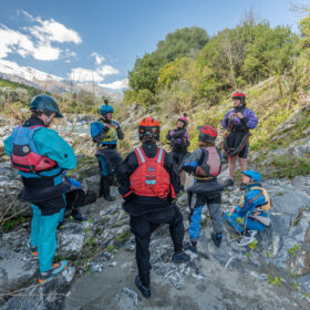 Group of hikers wearing helmets and life jackets stand on rocky canyon terrain, listening to a guide in purple gear.