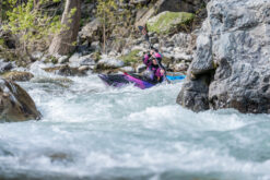 Person wearing a pink helmet paddles a purple kayak through fast whitewater between rocks.