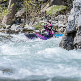Person wearing a pink helmet paddles a purple kayak through fast whitewater between rocks.
