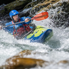 Kayaker wearing a blue helmet and red life jacket paddling a lime-green kayak through whitewater rapids with an orange paddle.