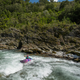 A person whitewater kayaking on a fast river with rocky banks and dense green forest under a blue sky