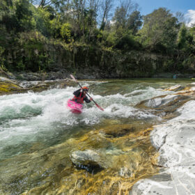 Paddler in a bright pink kayak navigating fast whitewater on a rocky river, wearing a red life jacket and helmet.