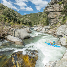 Kayaker paddling a blue kayak through swift whitewater between rocky cliffs in a canyon.