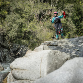 Person in helmet and protective gear standing on a rock by a fast-flowing river in a rocky canyon, raising arms in celebration.