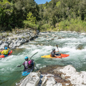 Group of kayakers navigating fast, rocky river with forested banks and rapids.