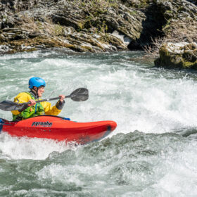 Person wearing a blue helmet paddling a red kayak through whitewater rapids in a rocky river scenery.