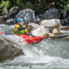 Person wearing a blue helmet paddles a red kayak through whitewater rapids beneath rocky surroundings.