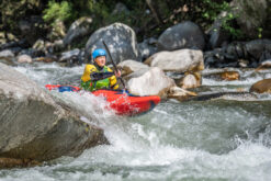 Person wearing a blue helmet paddles a red kayak through whitewater rapids beneath rocky surroundings.