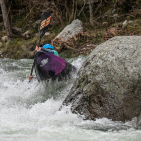 Kayaker in a blue helmet navigates whitewater around large rocks in a purple kayak.