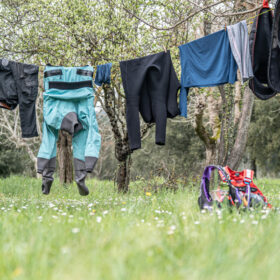 Clothesline between trees with drying outdoor gear in a grassy park; turquoise pants, jackets, and other clothing; a red backpack on the grass nearby.