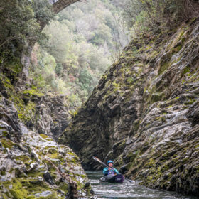 Kayaker in a turquoise jacket paddling through a narrow rocky river gorge beneath a stone arch bridge.