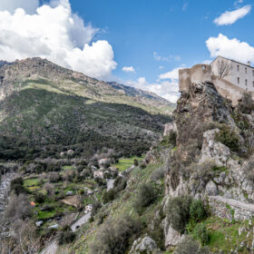 Medieval fortress perched on a rocky outcrop above a green valley and village.