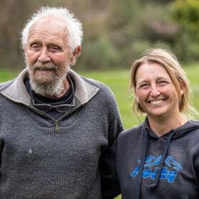Smiling older man with white hair and beard stands beside a woman in a dark hoodie in a green park.