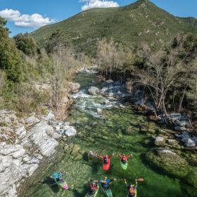 Six kayakers in bright gear leap into a clear green river, surrounded by rocky banks, trees, and a mountainside in the distance.
